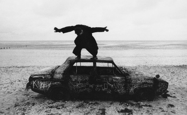 Man standing on a burnt-out car, covered in graffiti, on a beach